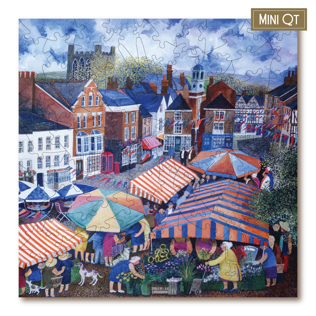 A completed jigsaw puzzle featuring a colorful village market scene with striped market tents, people, buildings, and a church tower under a cloudy sky. The puzzle is shown at a slight angle with a small gold label in the top-right corner that reads MINI QT.