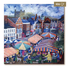 A completed jigsaw puzzle featuring a colorful village market scene with striped market tents, people, buildings, and a church tower under a cloudy sky. The puzzle is shown at a slight angle with a small gold label in the top-right corner that reads MINI QT.