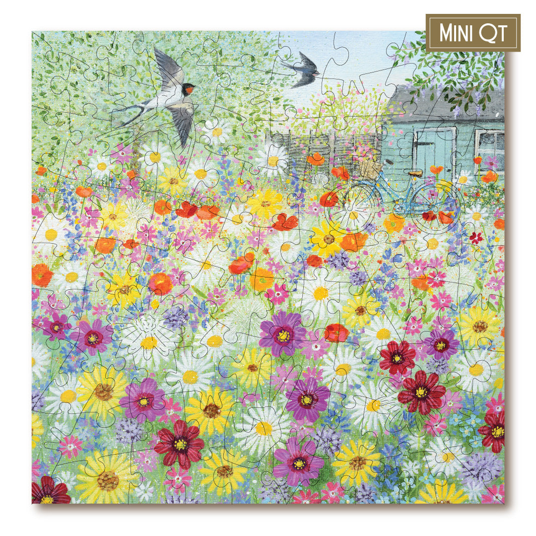 A completed jigsaw puzzle featuring a colorful wildflower meadow with blooming flowers, two flying birds, a bicycle, and a small house. The puzzle is shown at a slight angle with a brown label in the top right corner that reads 'MINI QT'.